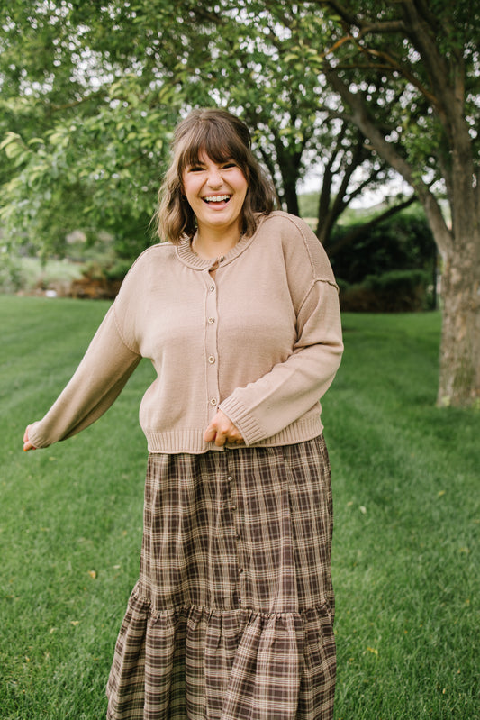 A woman smiling outdoors on a grassy lawn with trees in the background, wearing a beige button-front cardigan and a brown plaid maxi skirt.