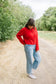 A woman wearing a bright red button-up cardigan, light-wash wide-leg jeans, and white sneakers stands on a dirt path surrounded by green trees.
