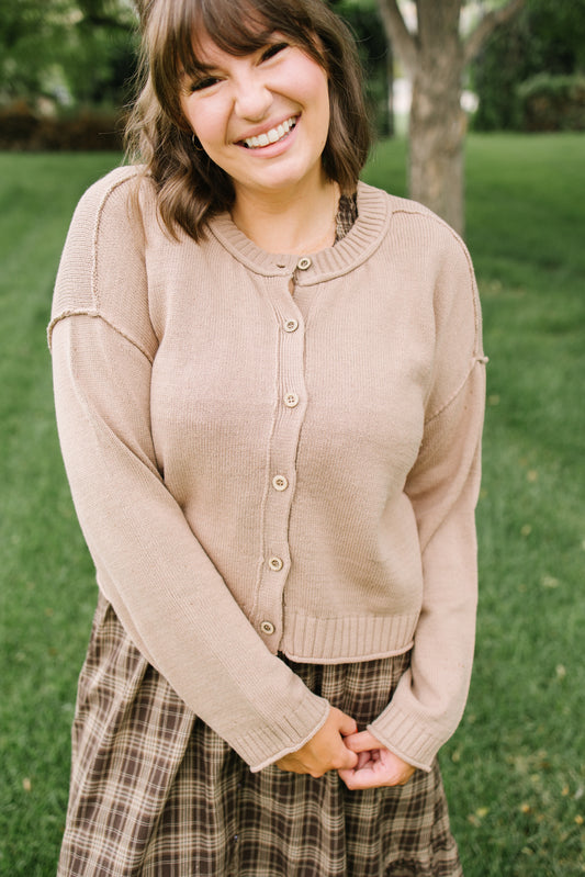 A smiling woman outdoors wearing a light beige knit cardigan with button-front closure and a brown plaid skirt, standing on a grassy lawn with trees in the background.