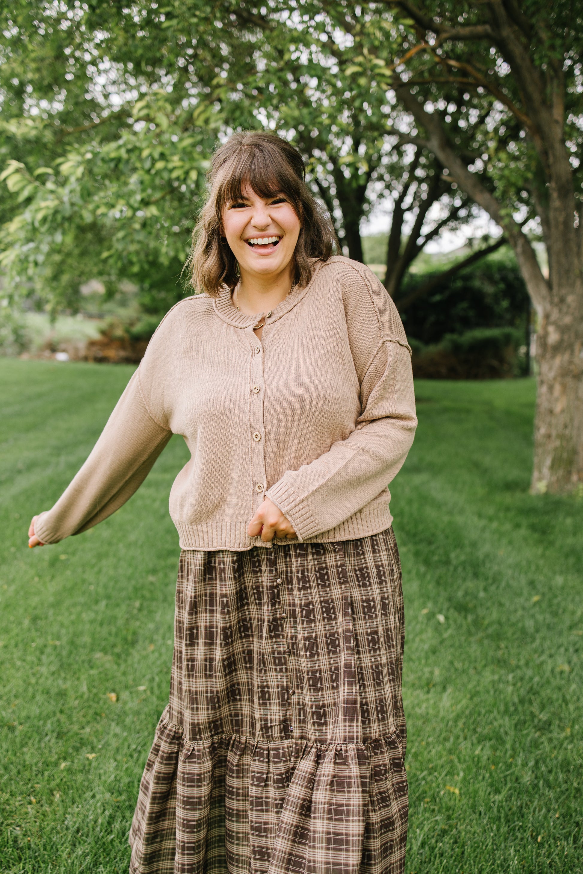 A woman smiling outdoors on a grassy lawn with trees in the background, wearing a beige button-front cardigan and a brown plaid maxi skirt.