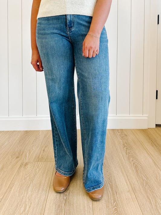 Person wearing blue jeans and brown shoes standing on a wooden floor.