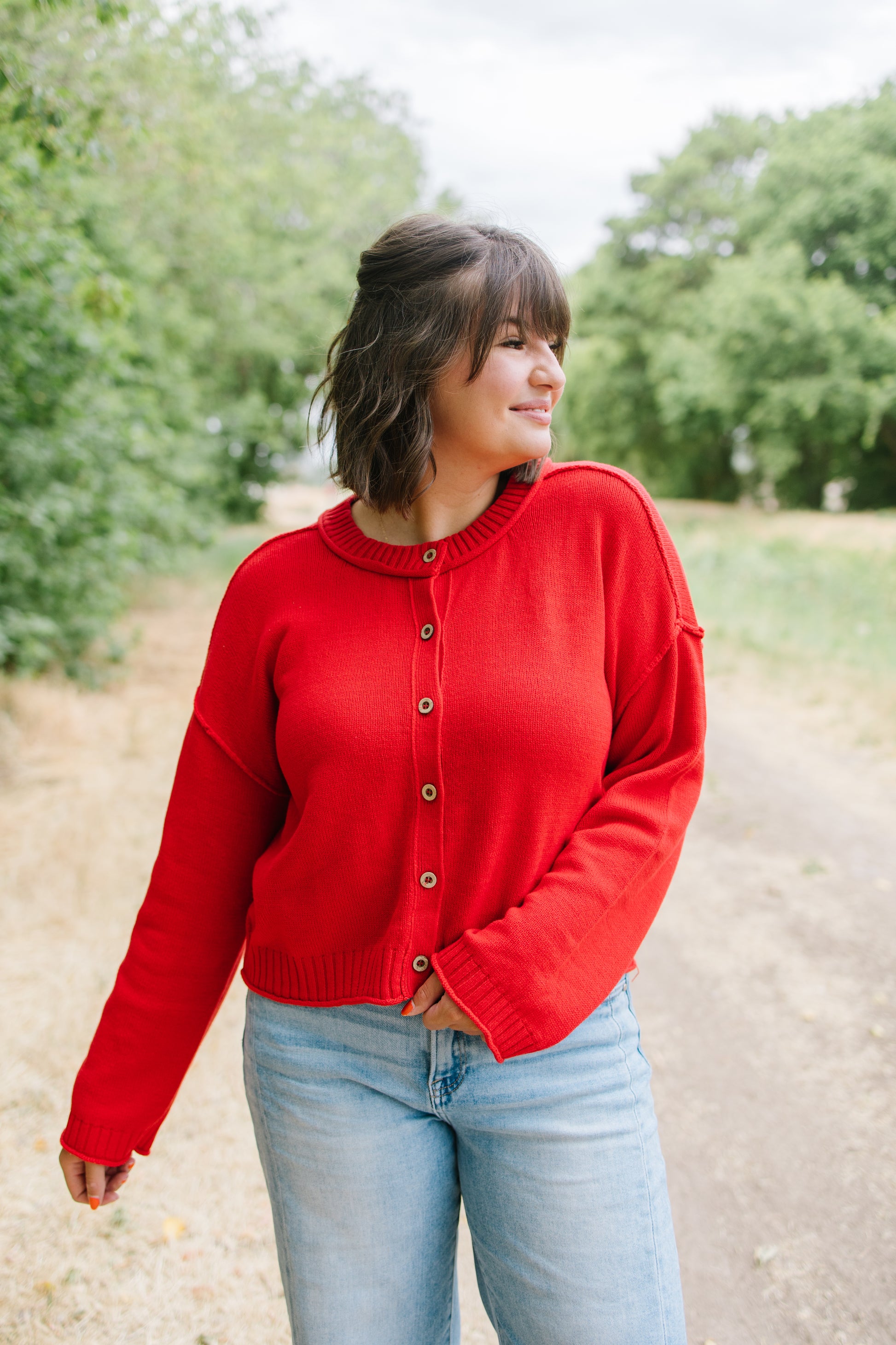 A woman wearing a bright red button-up cardigan sweater and light blue jeans, standing outdoors on a dirt path with trees in the background.