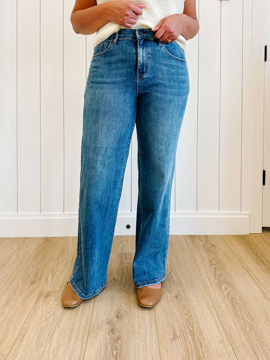 Person wearing blue jeans standing on a wooden floor with a white paneled wall in the background