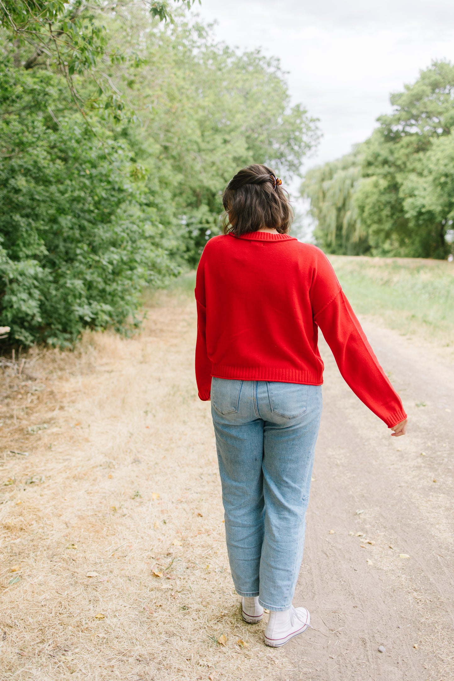 A woman seen from behind walking on a dirt path outdoors, wearing a bright red knit sweater, light blue jeans, and white sneakers.