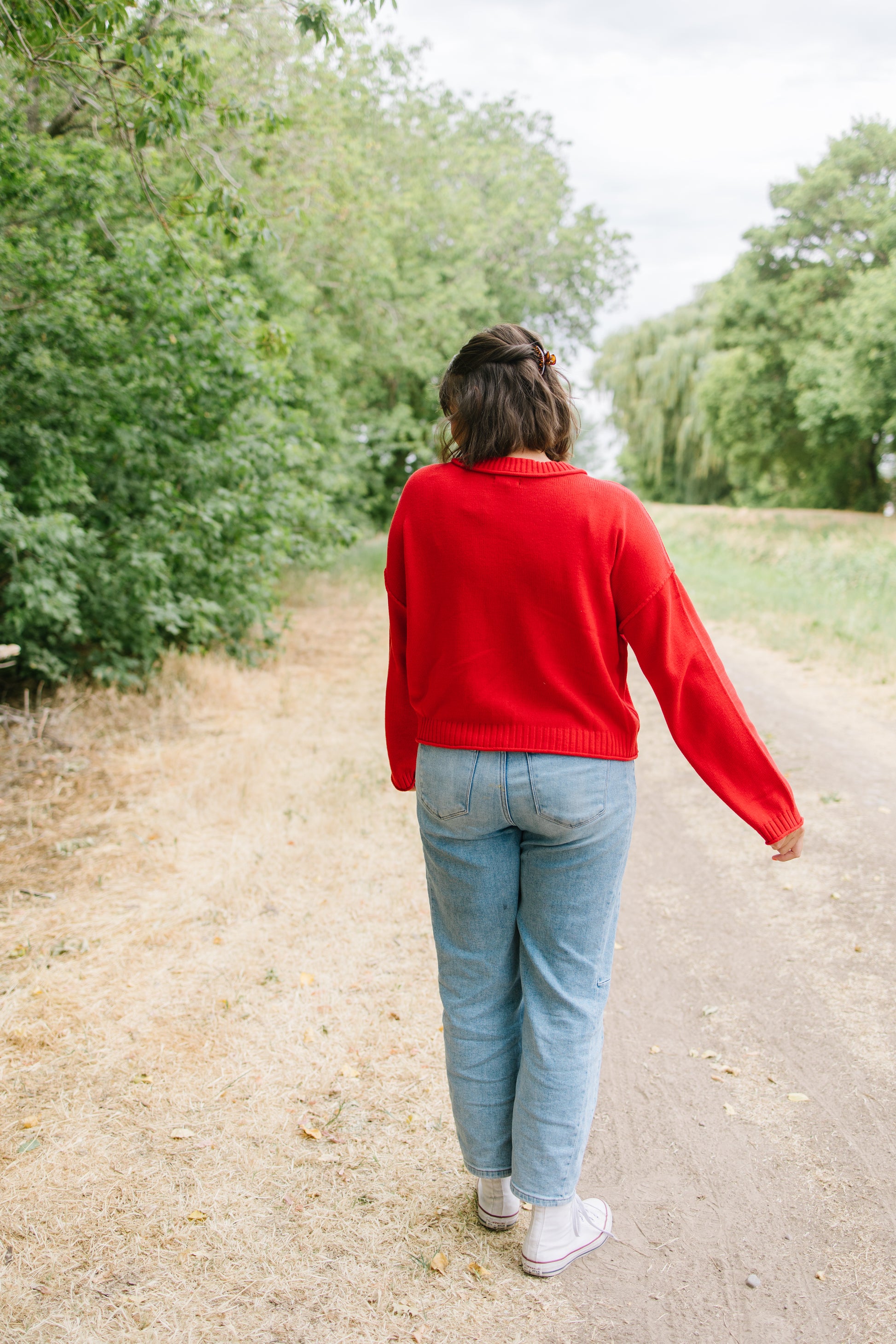 A woman seen from behind walking on a dirt path outdoors, wearing a bright red knit sweater, light blue jeans, and white sneakers.