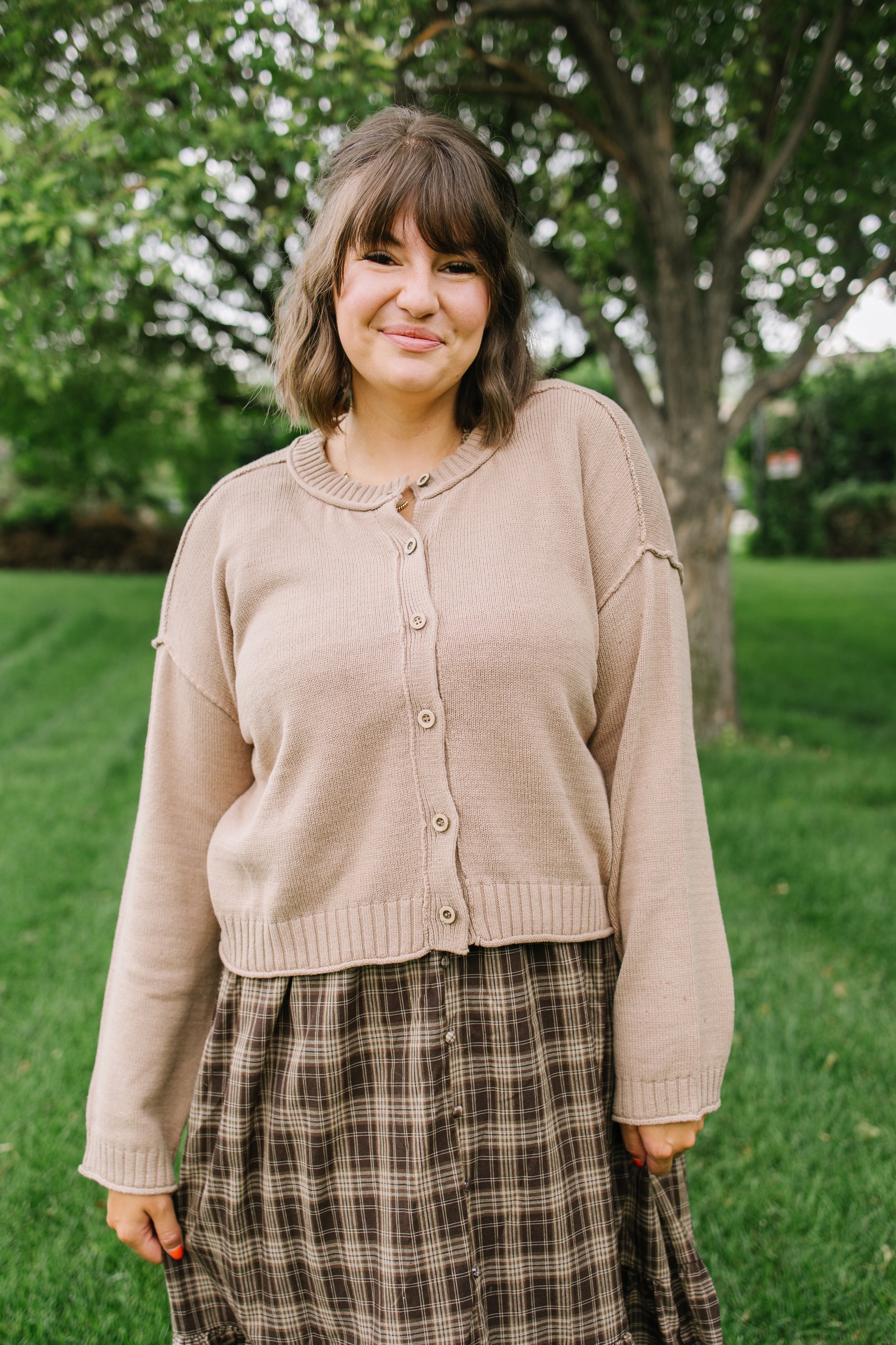 A woman wearing a beige button-up cardigan and a brown plaid skirt, standing outdoors in a grassy park area with trees in the background.