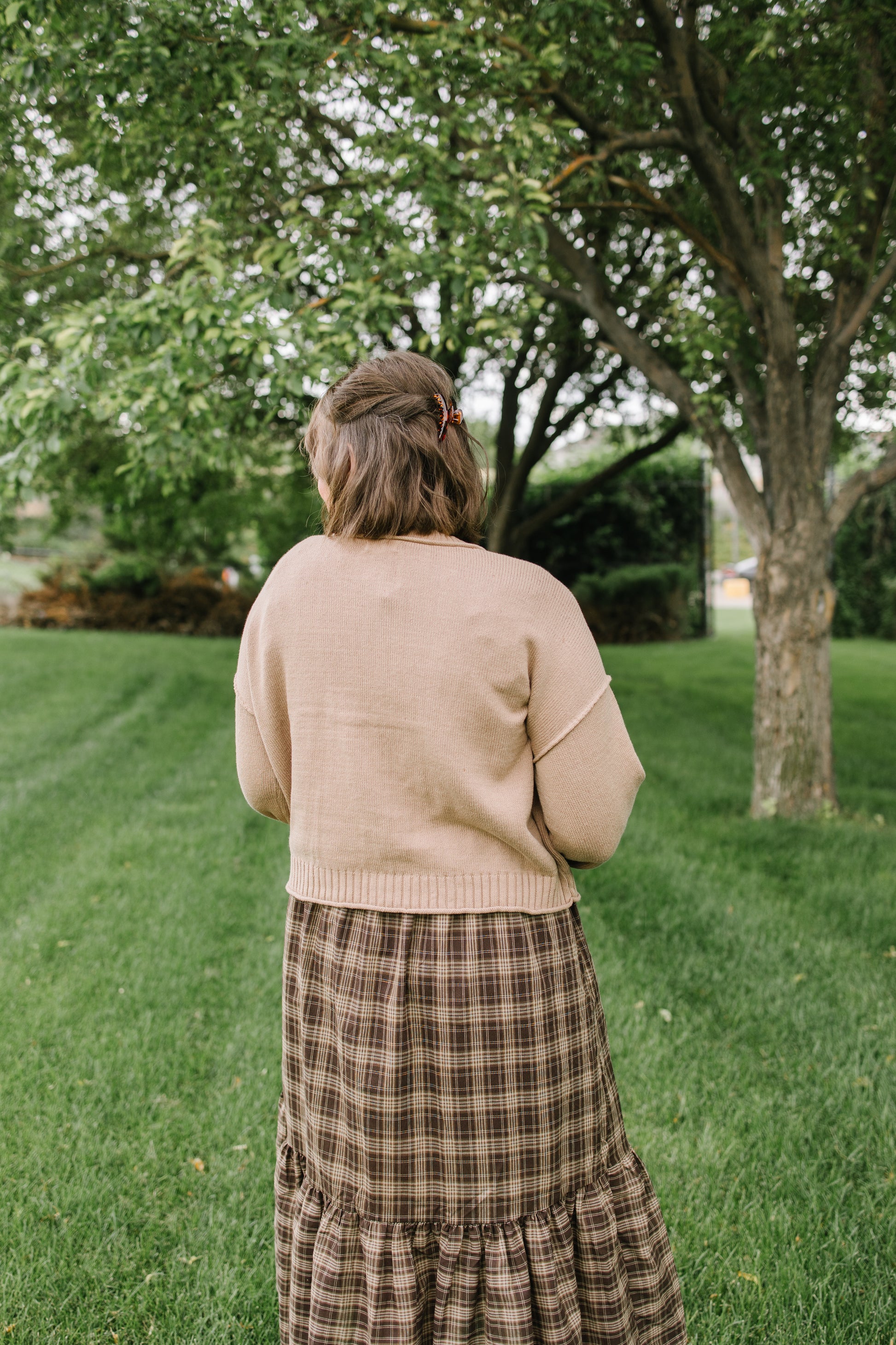 A woman viewed from behind wearing a beige knit cardigan and a brown plaid tiered skirt, standing on a grassy lawn with a large tree in the background.