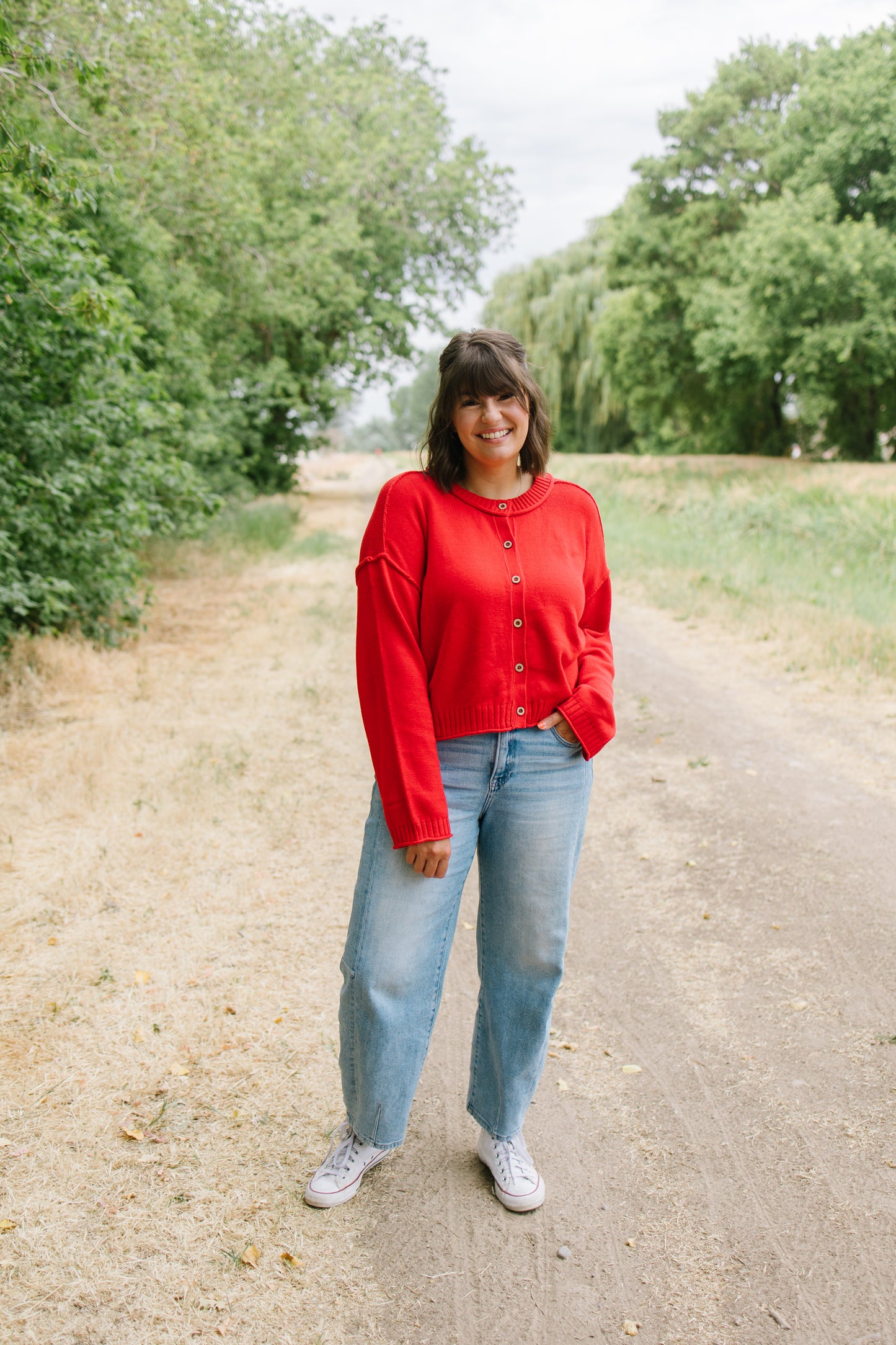 A woman stands on a dirt path outdoors wearing a bright red button-up cardigan, light blue jeans, and white sneakers.