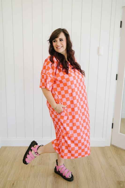 Woman wearing a pink and orange checkered dress standing in a room with white paneled walls.