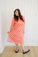 Woman wearing a bright orange and pink checkered dress with black sandals against a white paneled wall.