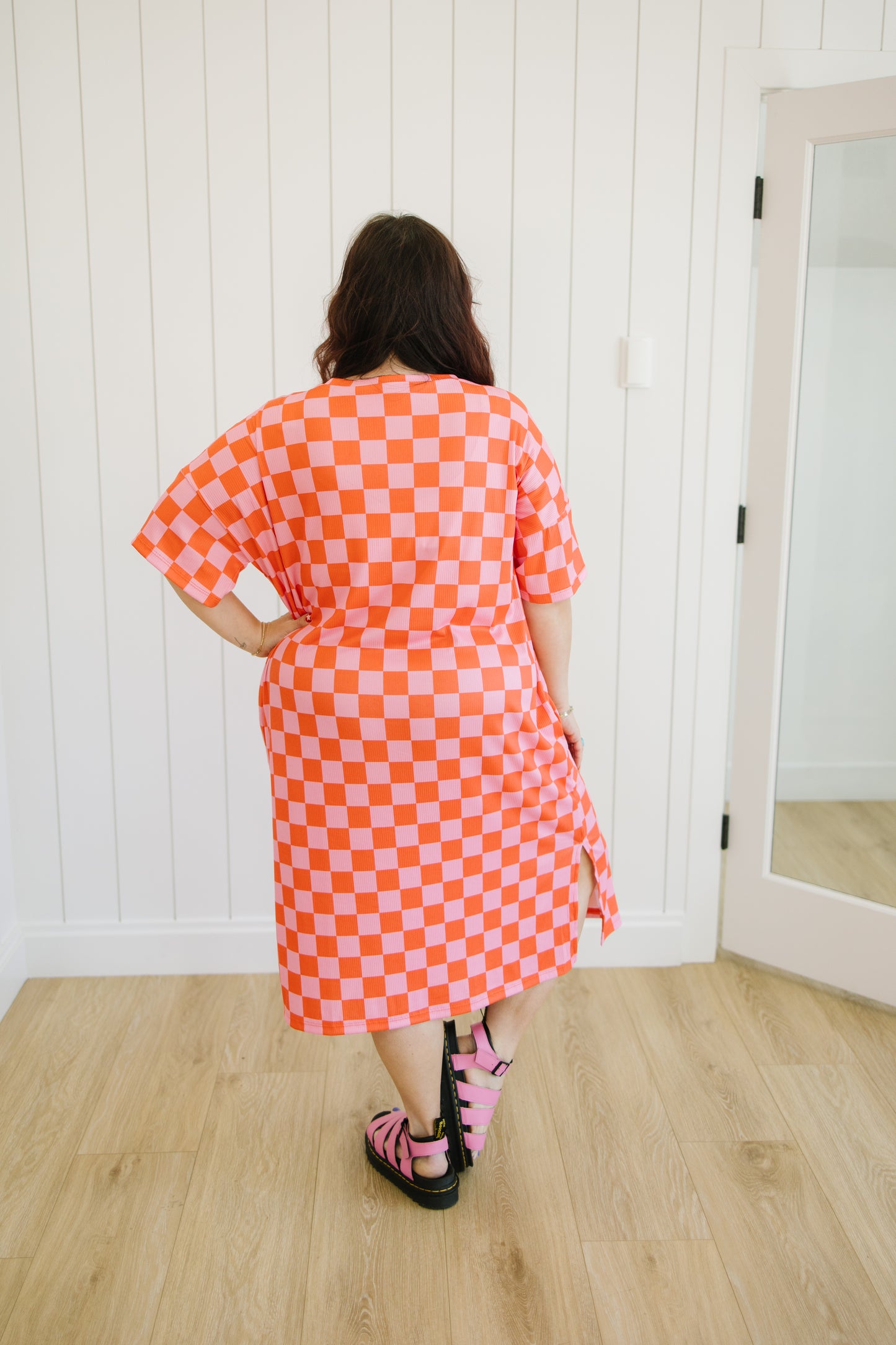 Person wearing a bright orange and white checkered dress standing in a room with wooden flooring and white walls.