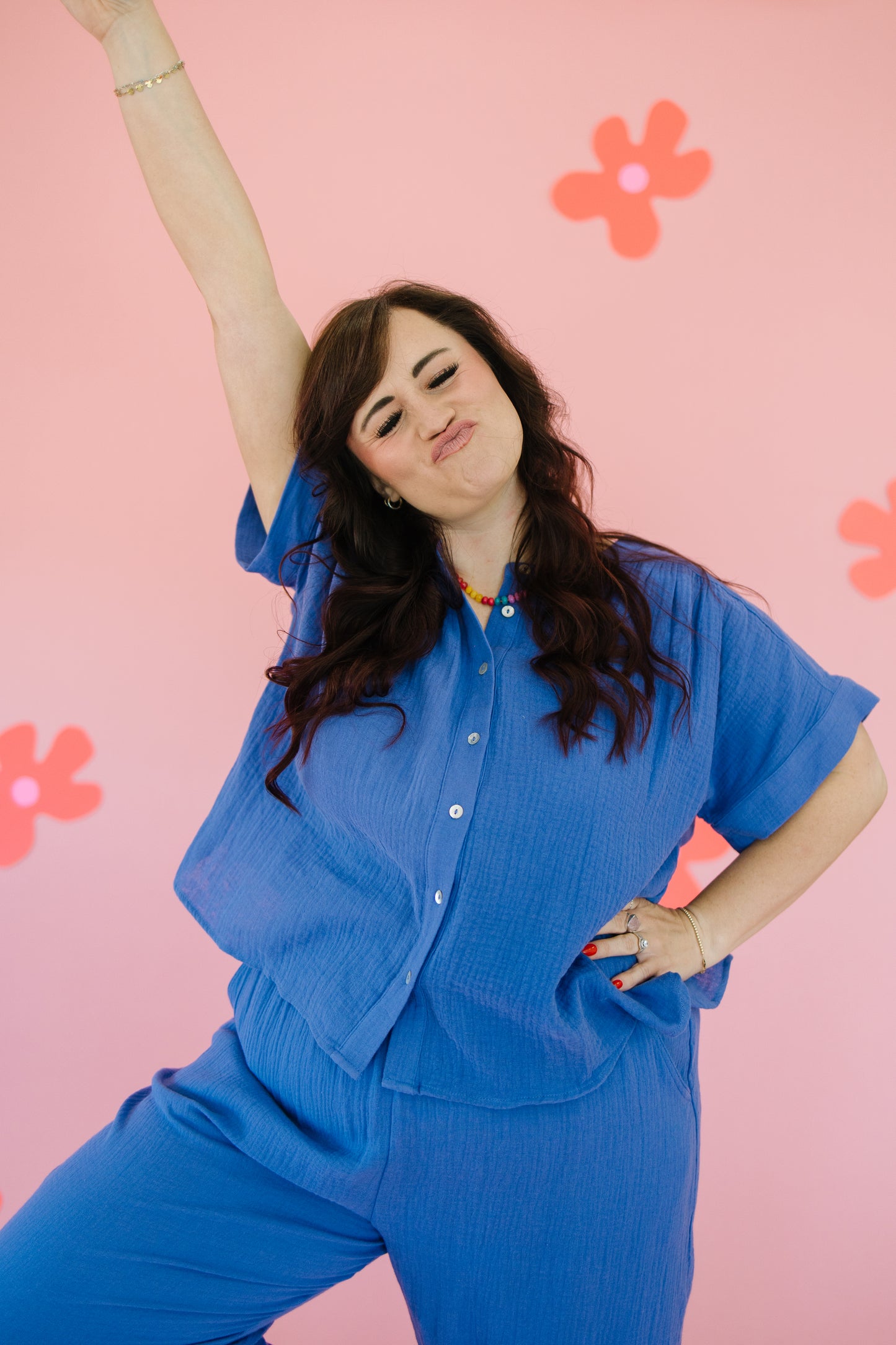 Woman in a blue outfit posing with arms raised against a pink background with flowers.