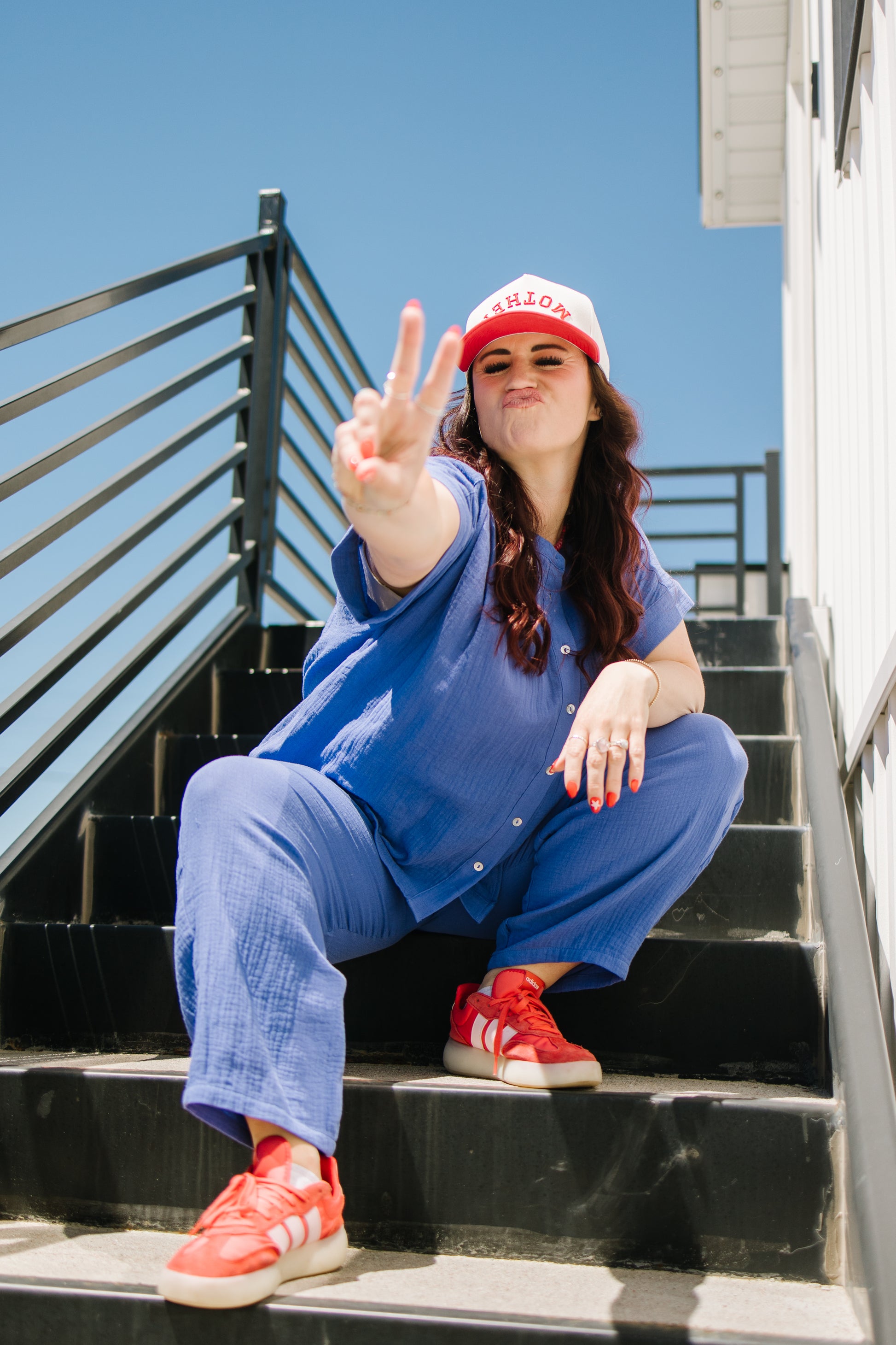 Person in blue jumpsuit and red sneakers making a peace sign on black steps with clear blue sky.