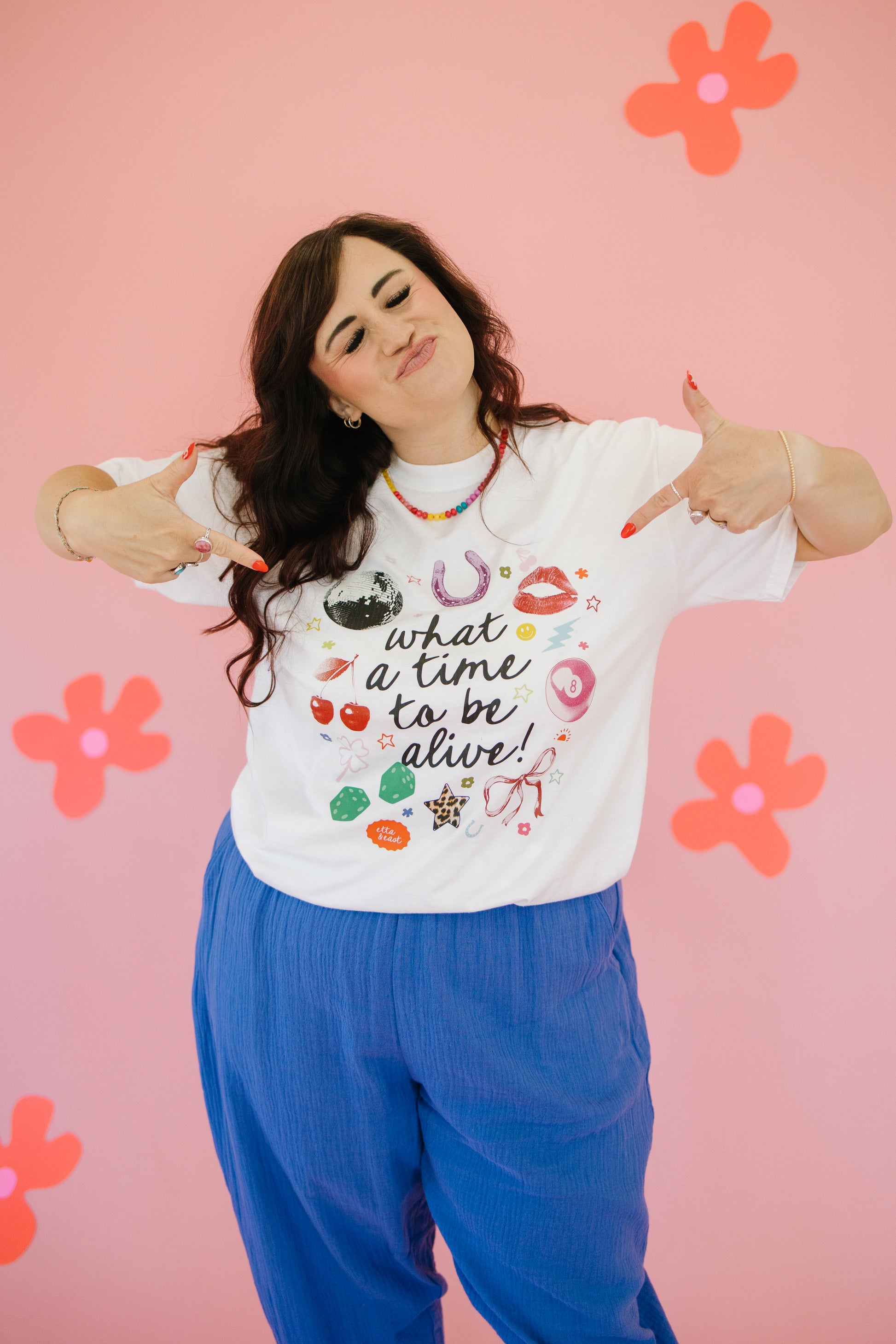 Person wearing a white t-shirt with colorful graphics and text, standing against a pink background with flowers.