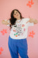 Person wearing a white t-shirt with colorful graphics and text, standing against a pink background with flowers.