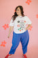 Woman wearing a white graphic t-shirt and blue pants on a pink background with flowers.