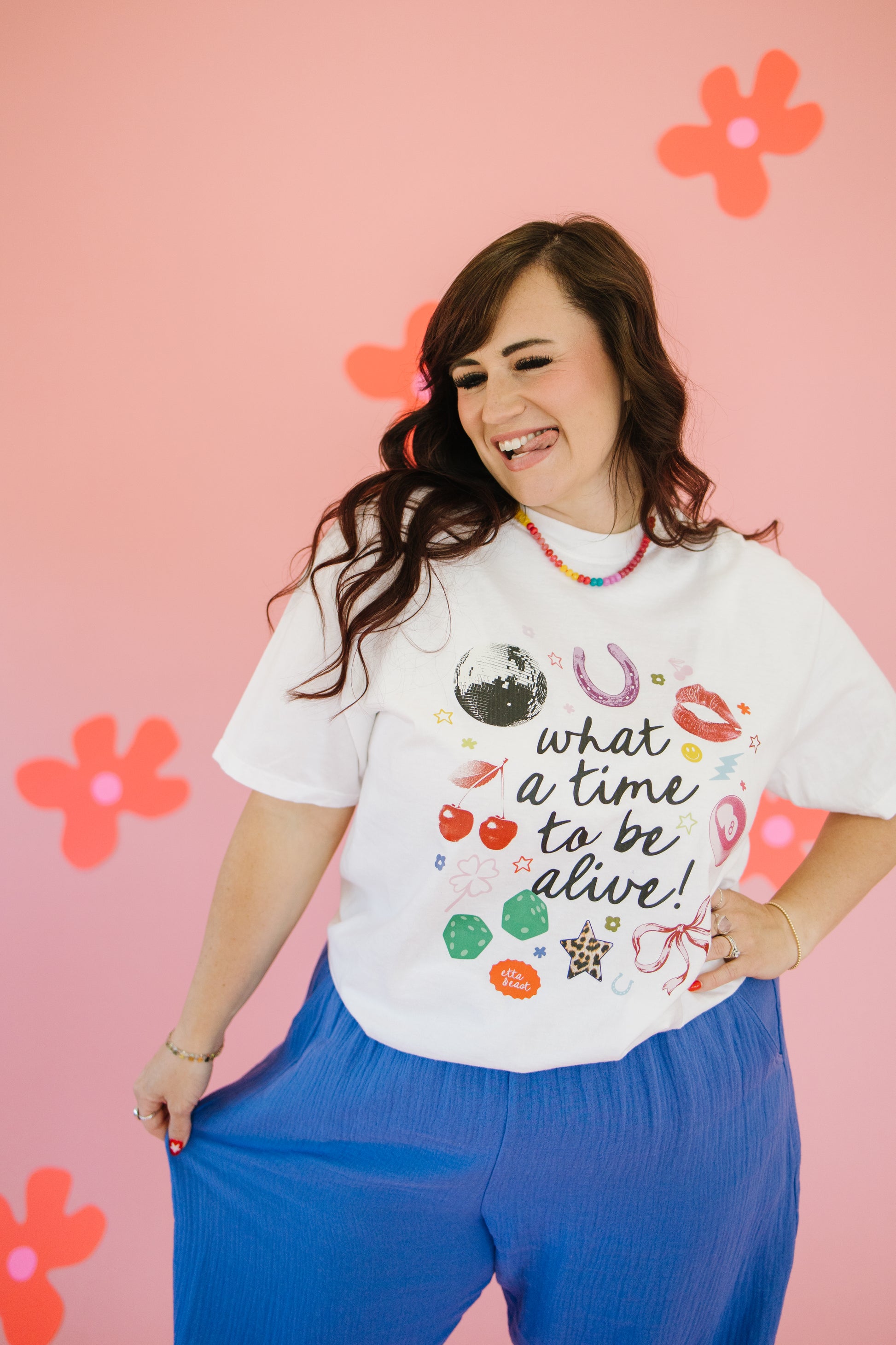 Woman wearing a white t-shirt with colorful graphics and text, standing against a pink background with flowers.