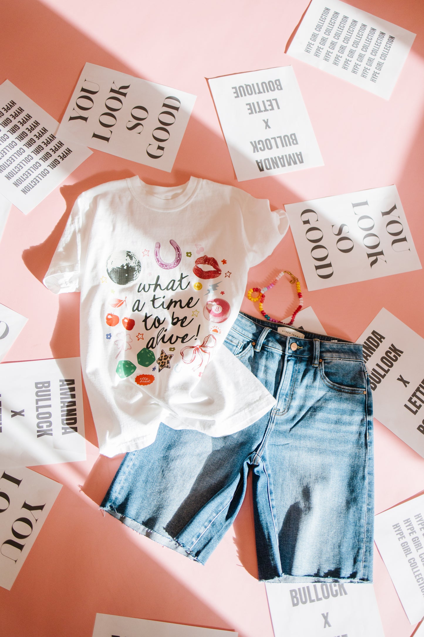 White t-shirt with colorful text and blue jeans on a pink surface with printed materials.
