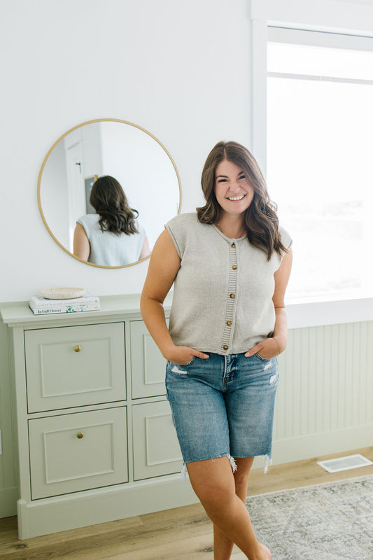 Woman standing in a room with a dresser and mirror, wearing a sleeveless sweater and denim shorts.