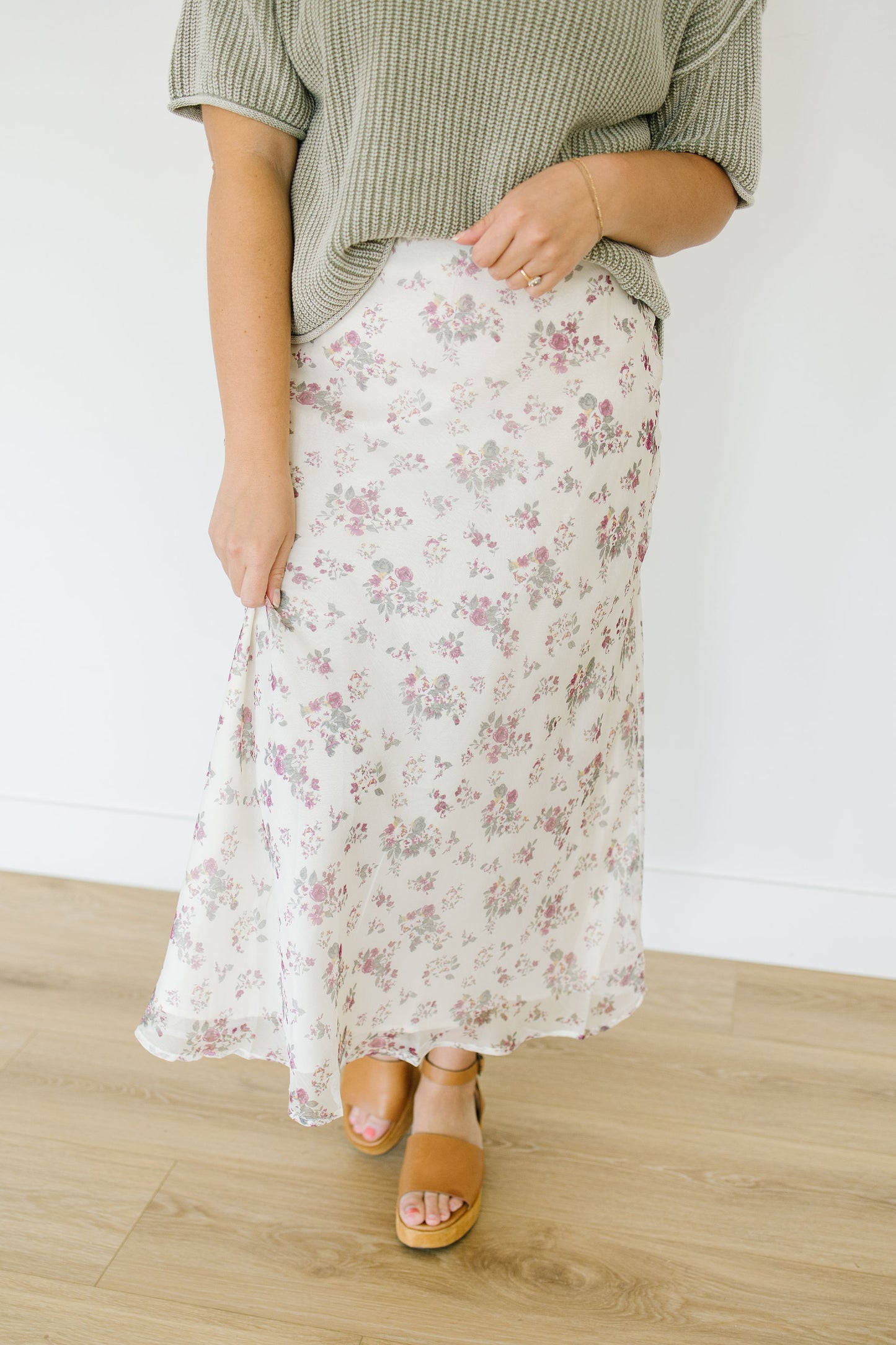 Person wearing a floral skirt and green top on a wooden floor with white wall background