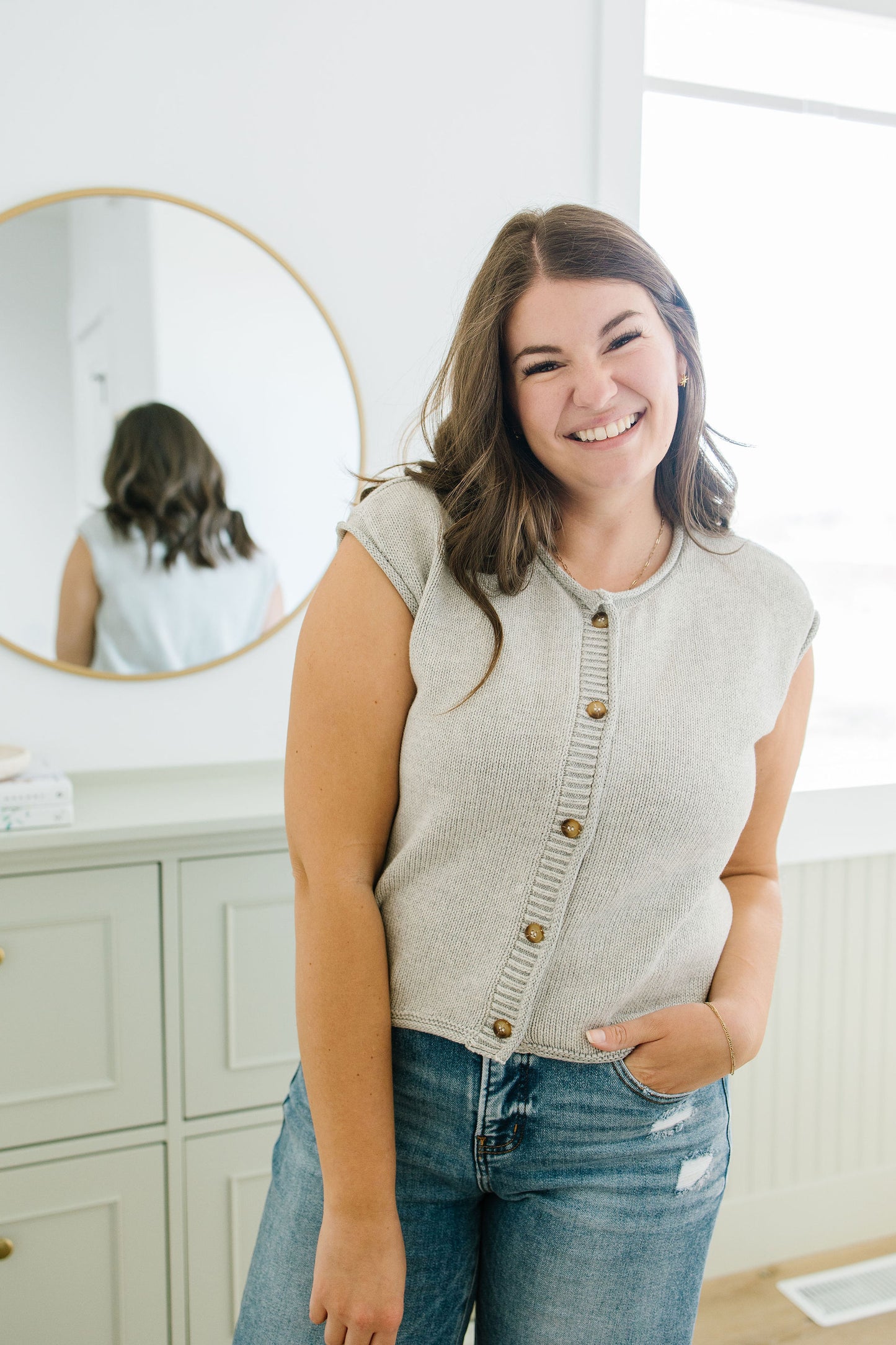 Woman wearing a light gray cardigan over a white shirt, standing in a room with a mirror and cabinets.