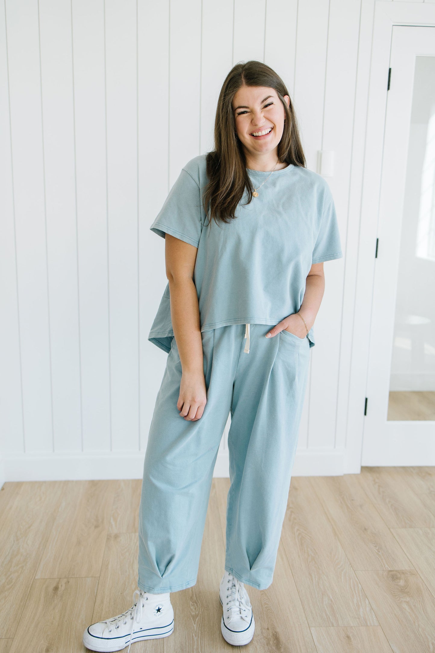 Woman wearing a light blue outfit standing in a room with wooden flooring and white walls.
