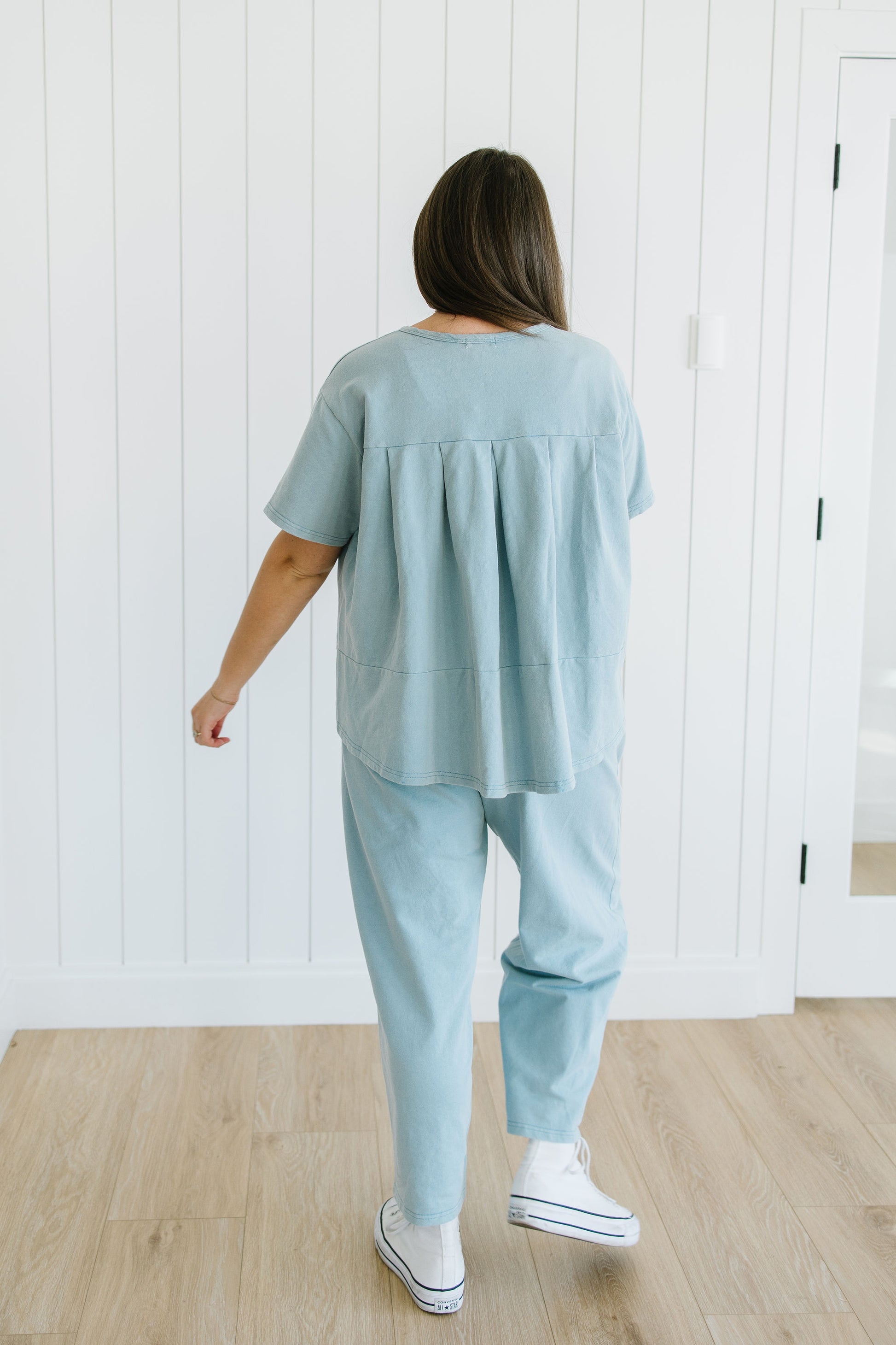 Person wearing a light blue outfit standing in a room with white walls and wooden flooring.