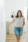 Woman wearing a sleeveless gray knit cardigan and denim shorts in a room with white walls and wooden flooring.