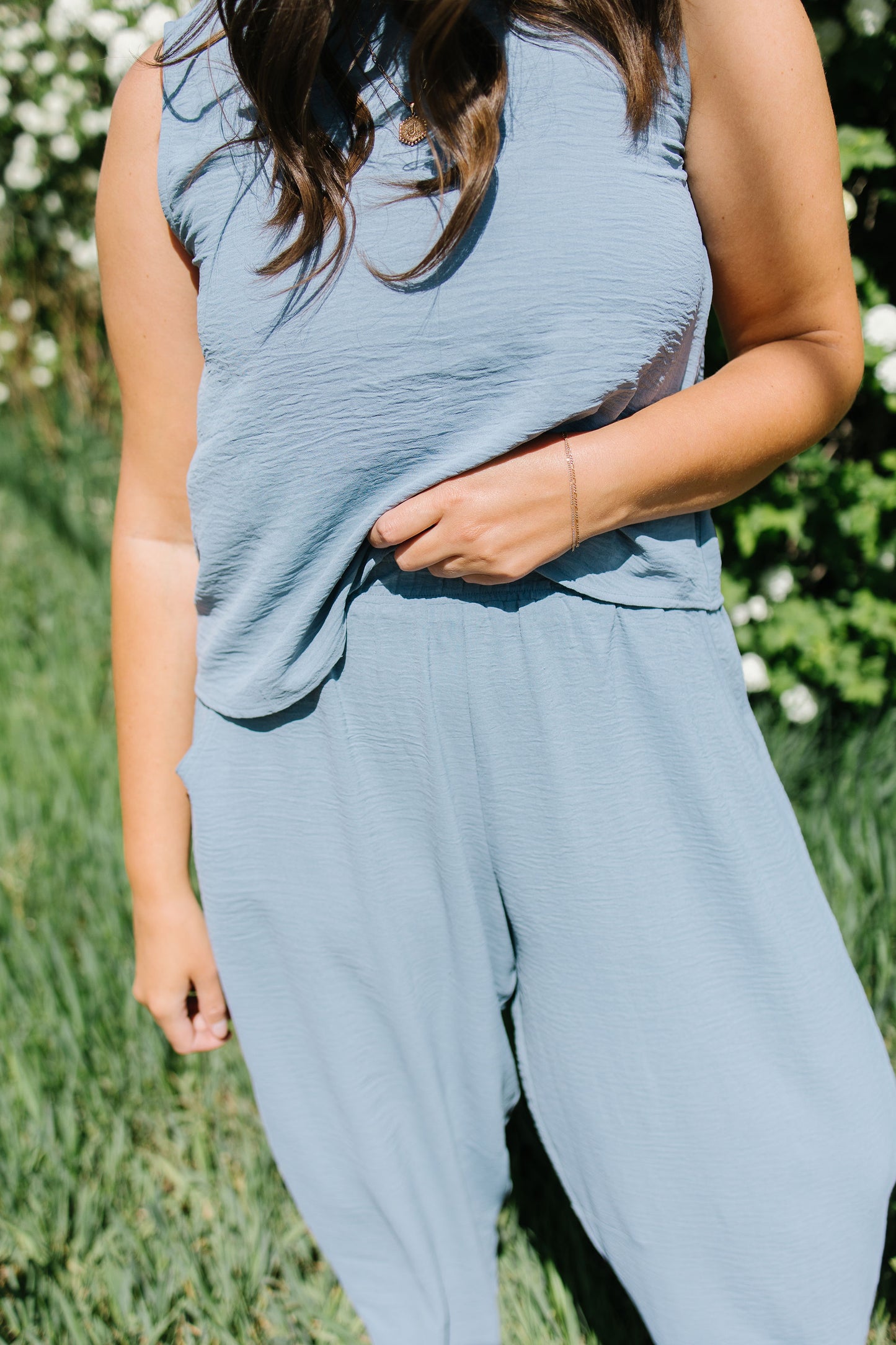 Person wearing a light blue sleeveless top and pants in an outdoor setting with greenery.