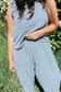 Person wearing a light blue sleeveless top and pants in an outdoor setting with greenery.