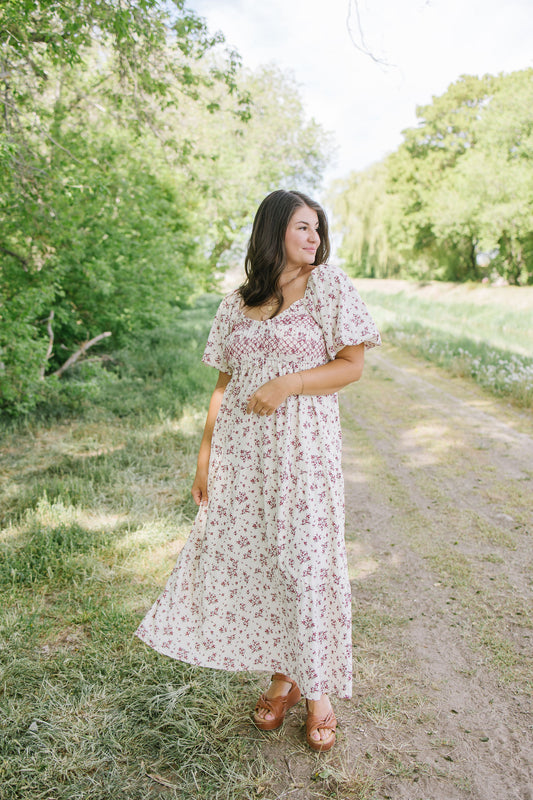 Woman in a floral dress standing on a dirt path with trees in the background