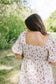 Woman in a floral dress standing in a natural setting with trees and greenery.