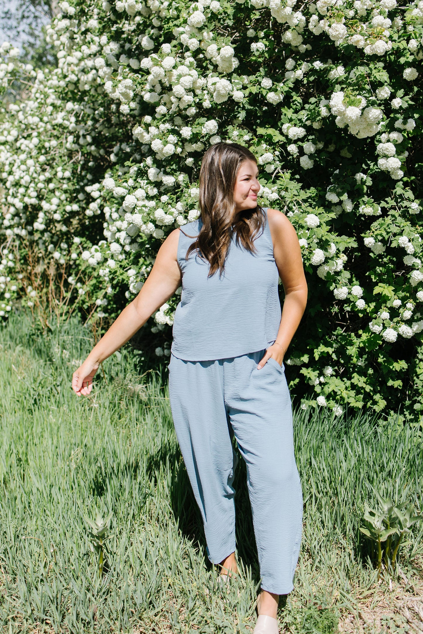 Woman in a light blue sleeveless top and pants standing in front of a white flowering bush.