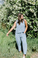 Woman in a light blue sleeveless top and pants standing in front of a white flowering bush.