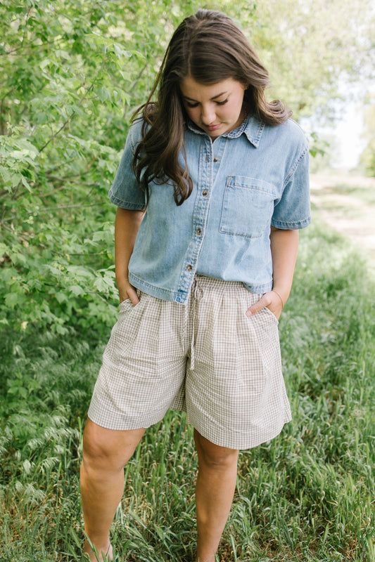 Woman wearing a denim shirt and plaid shorts standing in a grassy area with trees.