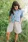 Woman wearing a denim shirt and plaid shorts standing in a grassy area with trees.