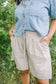 Person wearing a denim shirt and plaid shorts standing outdoors with greenery in the background