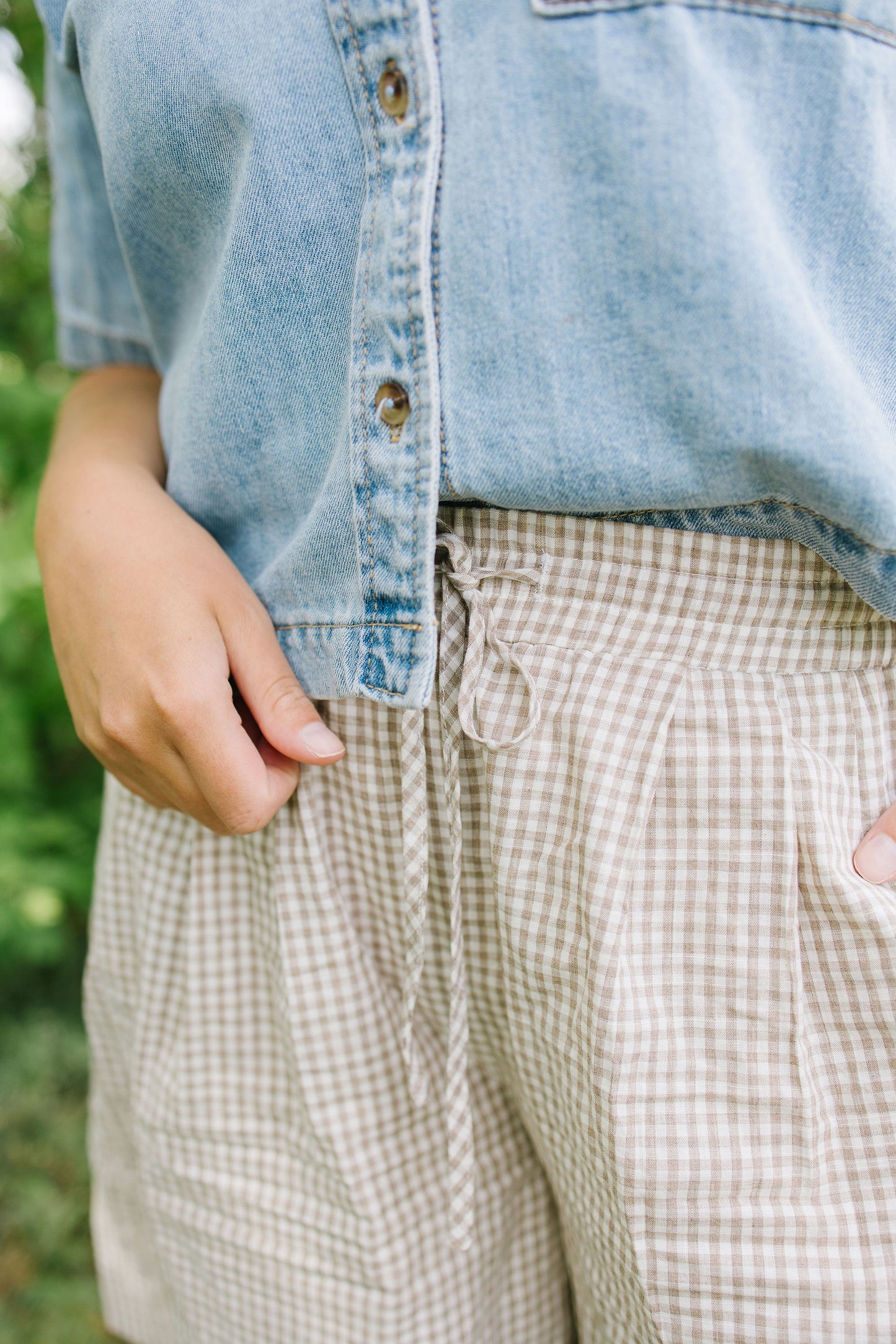 Person wearing a denim shirt and plaid pants with a blurred green background