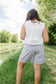 Person wearing a white sleeveless top and checkered shorts standing in a grassy area with trees and water in the background.