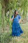 Woman in a blue dress standing in a natural setting with trees and grass.