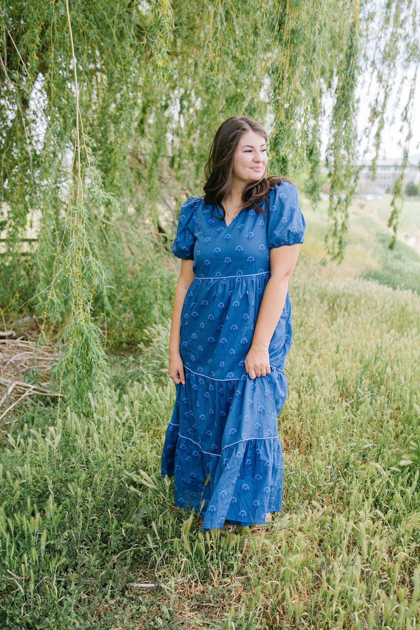 Woman in a blue dress standing in a field with a willow tree in the background
