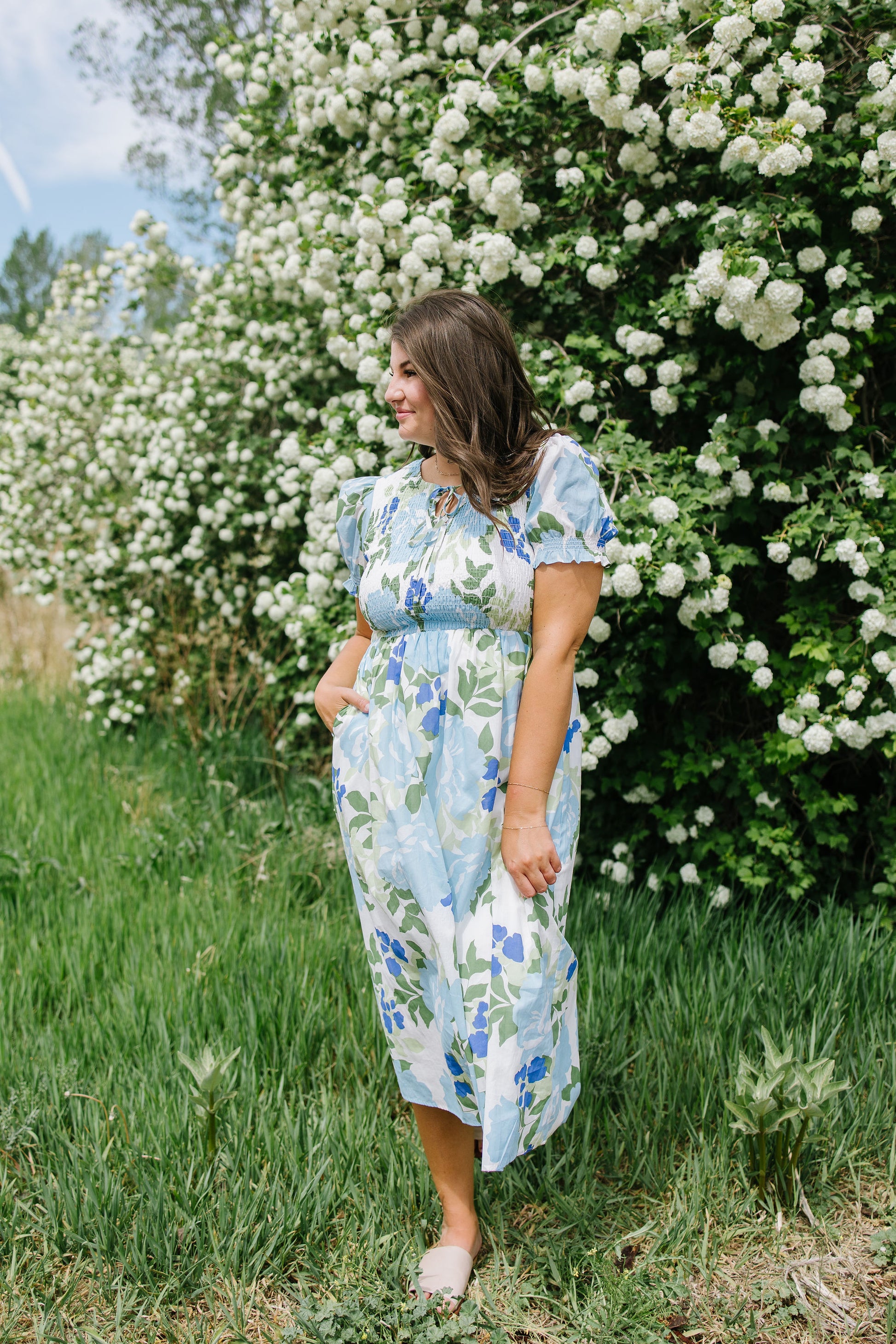 Woman in a floral dress standing in front of a white flowering bush