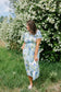 Woman in a floral dress standing in front of a white flowering bush