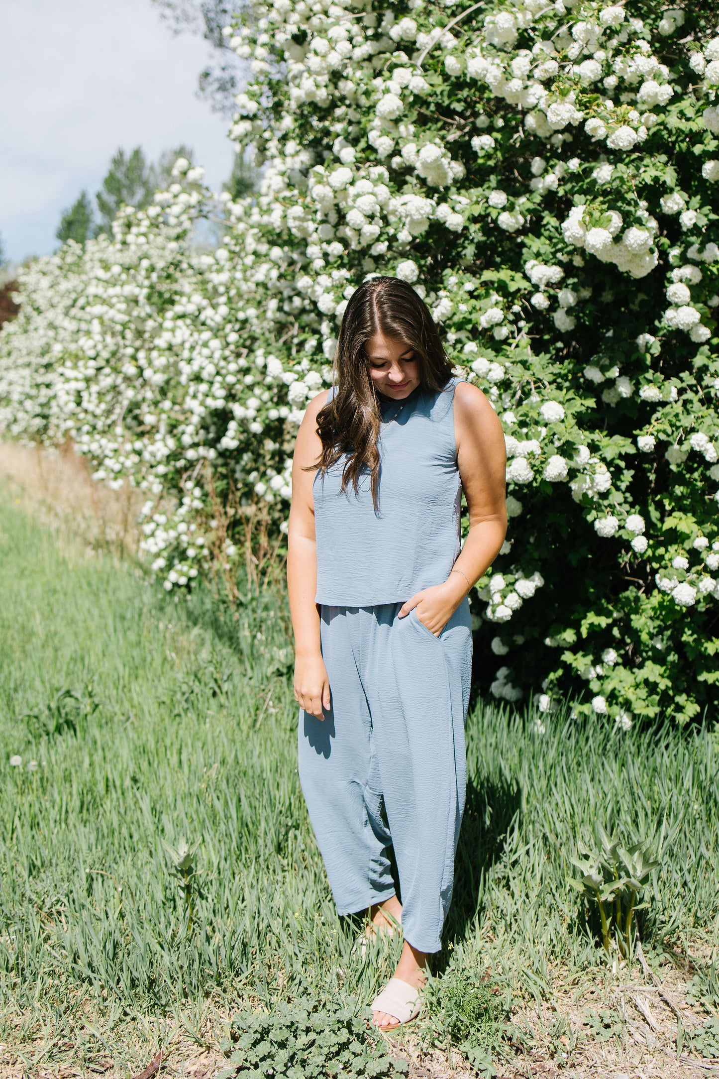 Woman in a light blue sleeveless top and harem pants standing in front of a white flowering bush.