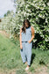 Woman in a light blue sleeveless top and harem pants standing in front of a white flowering bush.