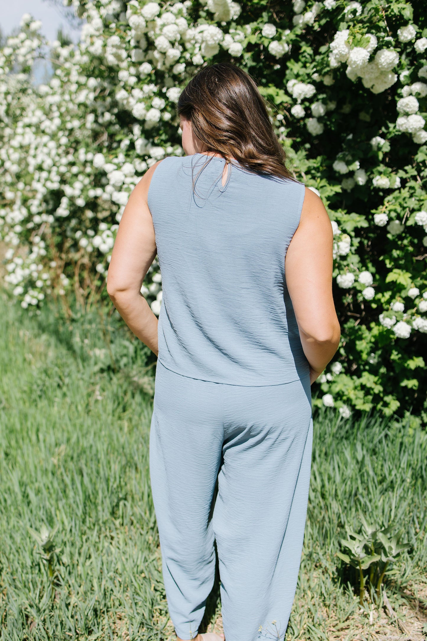 Person wearing a light blue sleeveless top and pants standing in front of a white flowering bush.