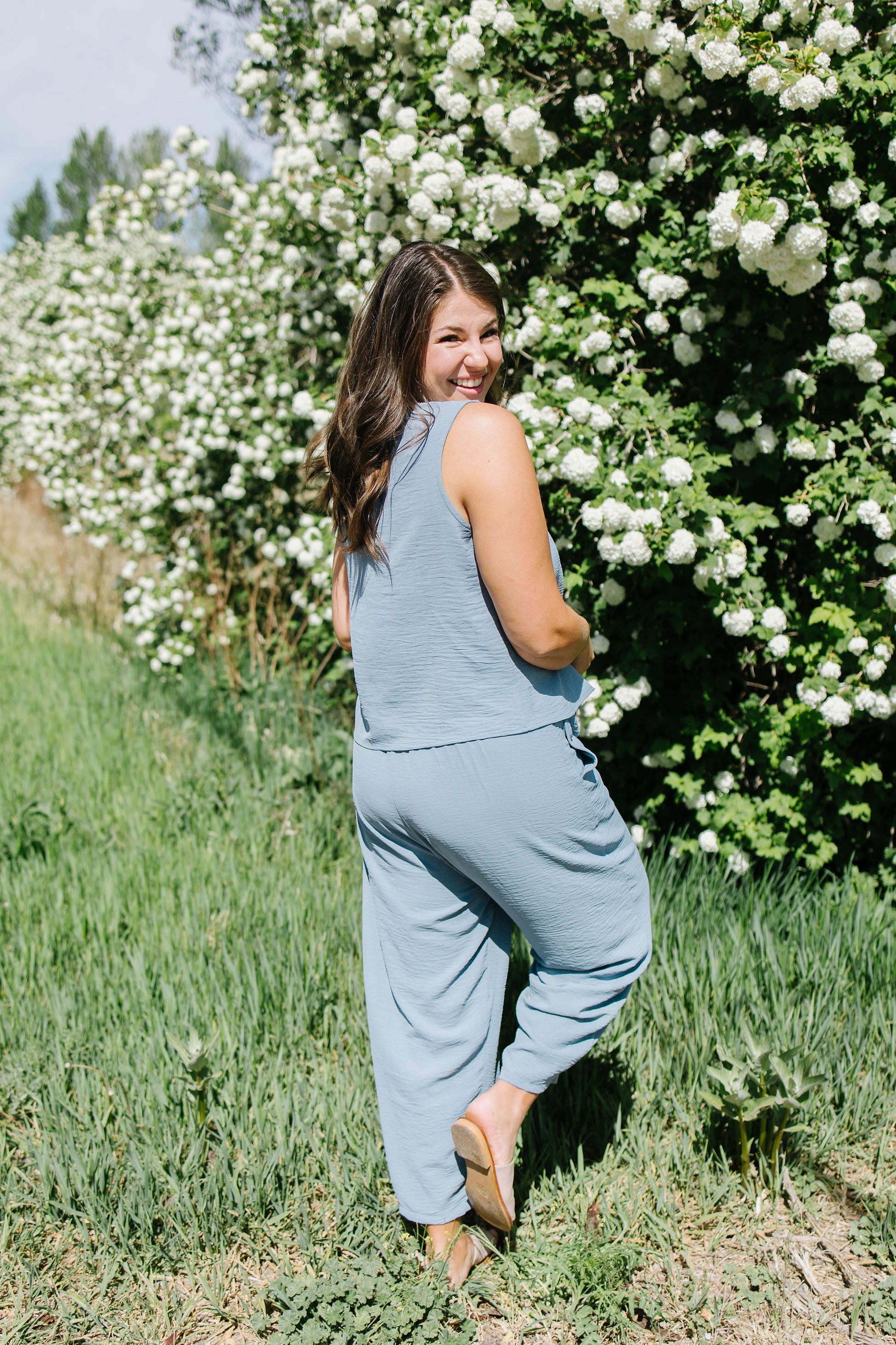 Woman in a light blue jumpsuit standing in front of white flowering bushes.