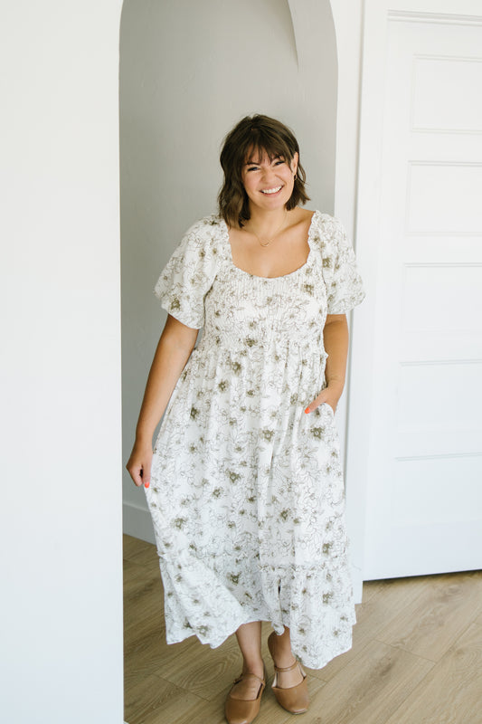 A woman wearing a white midi dress with a delicate brown floral pattern, standing in a bright room with white walls and light wood floors.