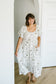 A woman wearing a white midi dress with a delicate brown floral pattern, standing in a bright room with white walls and light wood floors.