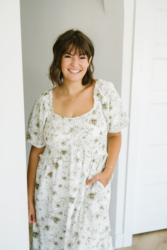 A woman smiling and wearing a white smocked dress with a muted green floral pattern, standing in a brightly lit indoor setting.